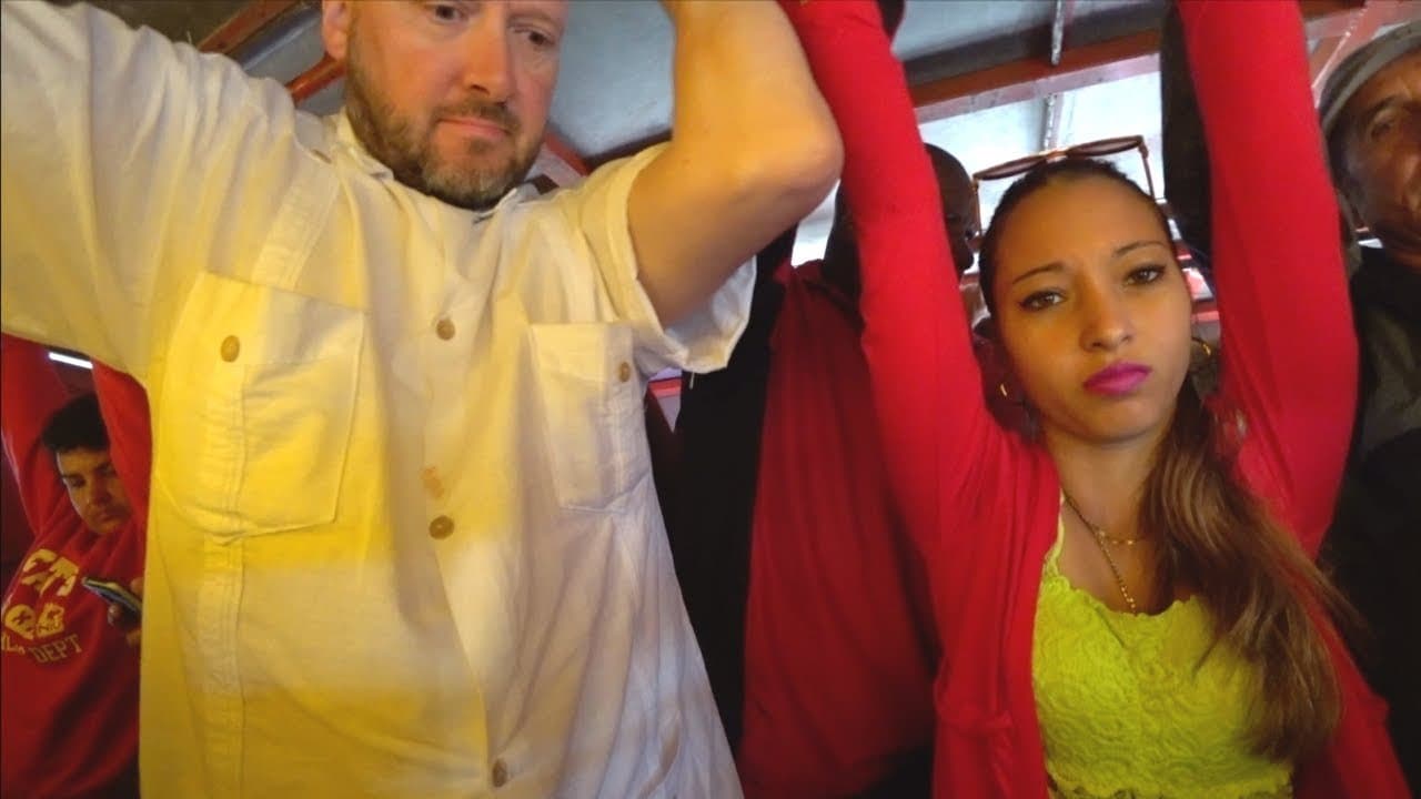 Benjamin Rich in a white short-sleeved shirt holding the overhead rail on a packed Cuban commuter bus, standing beside a young Cuban woman in a red cardigan and yellow lace top