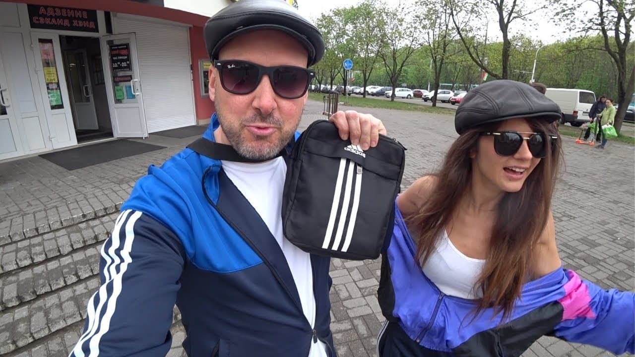 Benjamin Rich in a flat cap, sunglasses and a blue Adidas tracksuit top holding up a black Adidas shoulder bag, taking a selfie with his friend Alina, also in a flat cap and a retro tracksuit, outside a second-hand shop in Minsk, Belarus