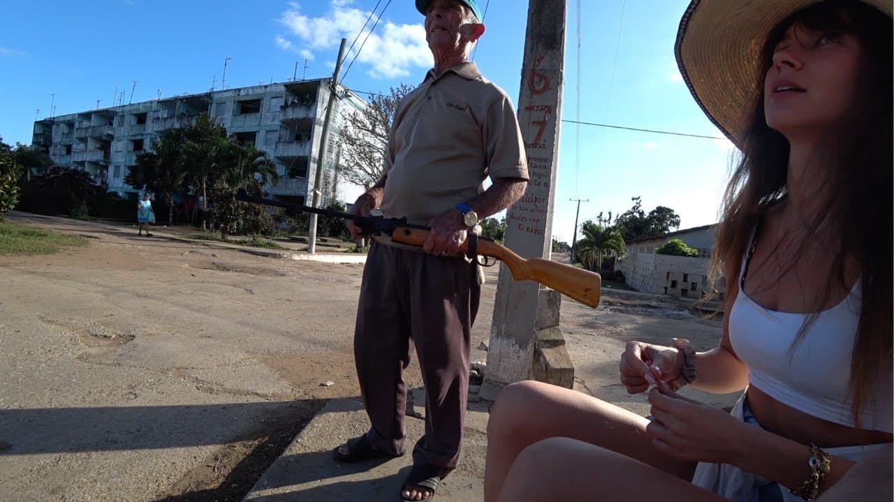 An elderly Cuban man in a cap and beige polo shirt holding an old rifle on a dusty small-town street corner, a weathered concrete apartment block and palm trees behind him, with Alina in a wide straw hat sitting in the foreground