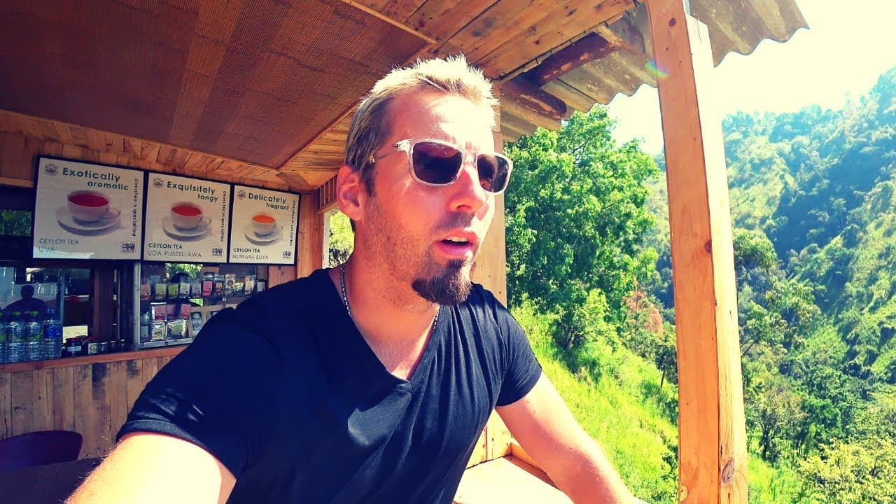 Harald Baldr squinting into the sun at a small wooden tea stall on the road to Ella, Sri Lanka, with framed Ceylon tea adverts on the wall behind him