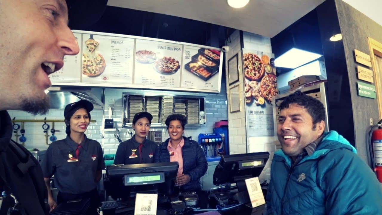 Kumar, in a teal puffer jacket, laughing at the counter of a Pizza Hut in Kathmandu while three smiling staff look on, with Harald's grinning face at the left edge of the frame