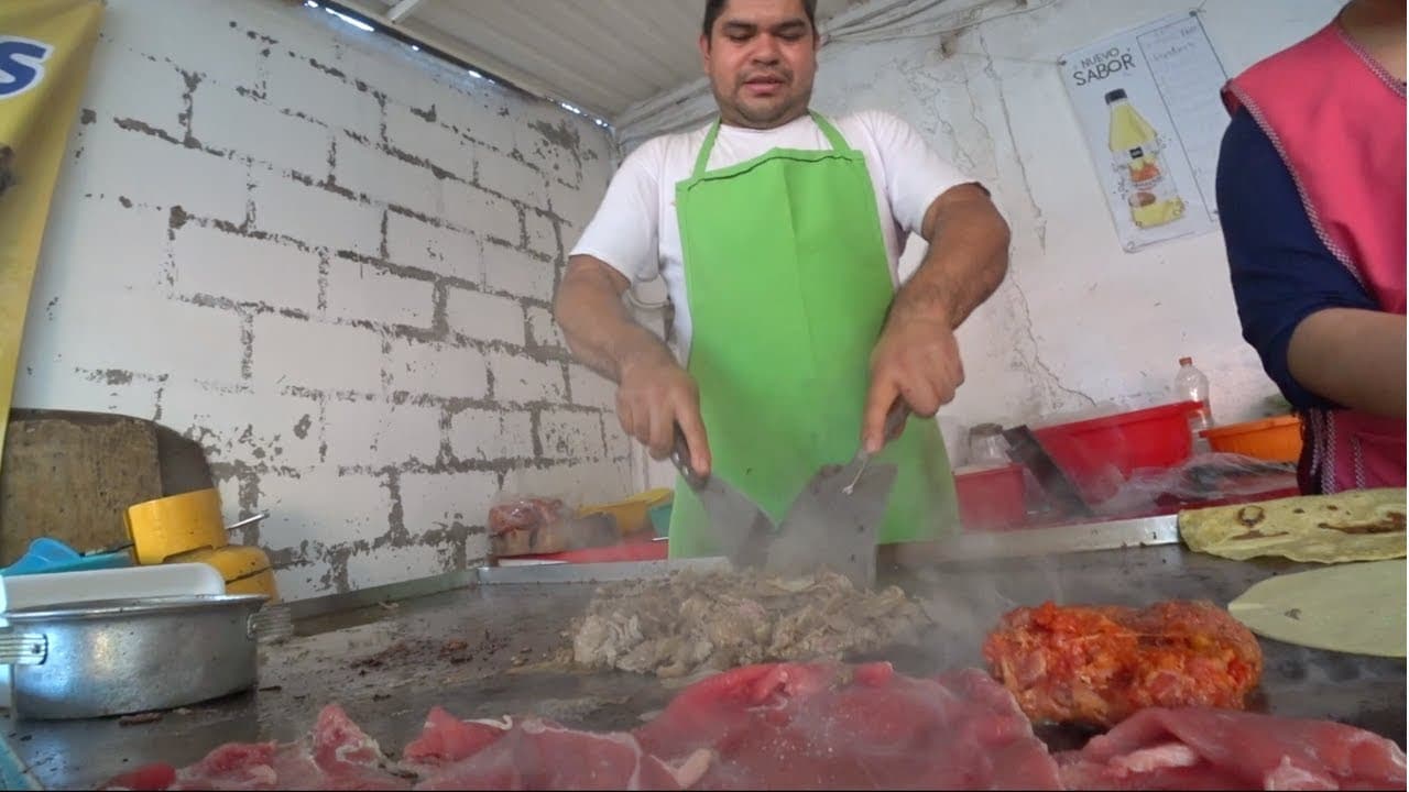 A taquero in a green apron chopping meat with two cleavers on a steaming flat-top grill at Los Super Tacos in Mexico City, with raw meat and tortillas in front