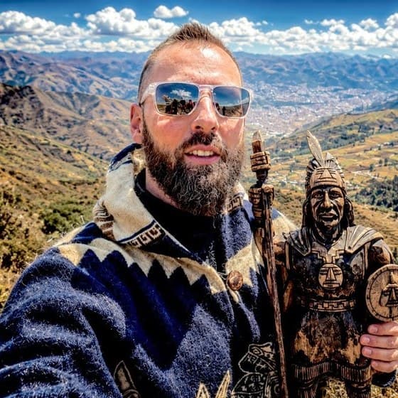 Harald Baldr, bearded, in mirrored sunglasses and a navy and cream Andean wool jacket, smiling in a selfie on a mountainside in Peru while holding a carved wooden statue of an Inca warrior, with a valley town far below