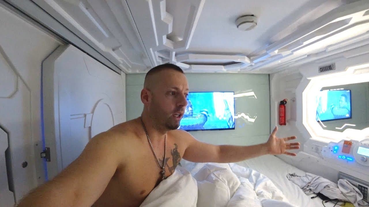 Harald Baldr, shirtless with a shaved head, sitting up in the white bed of a spaceship-style capsule hotel pod in Saigon, pointing at the wall TV and the glowing control panel