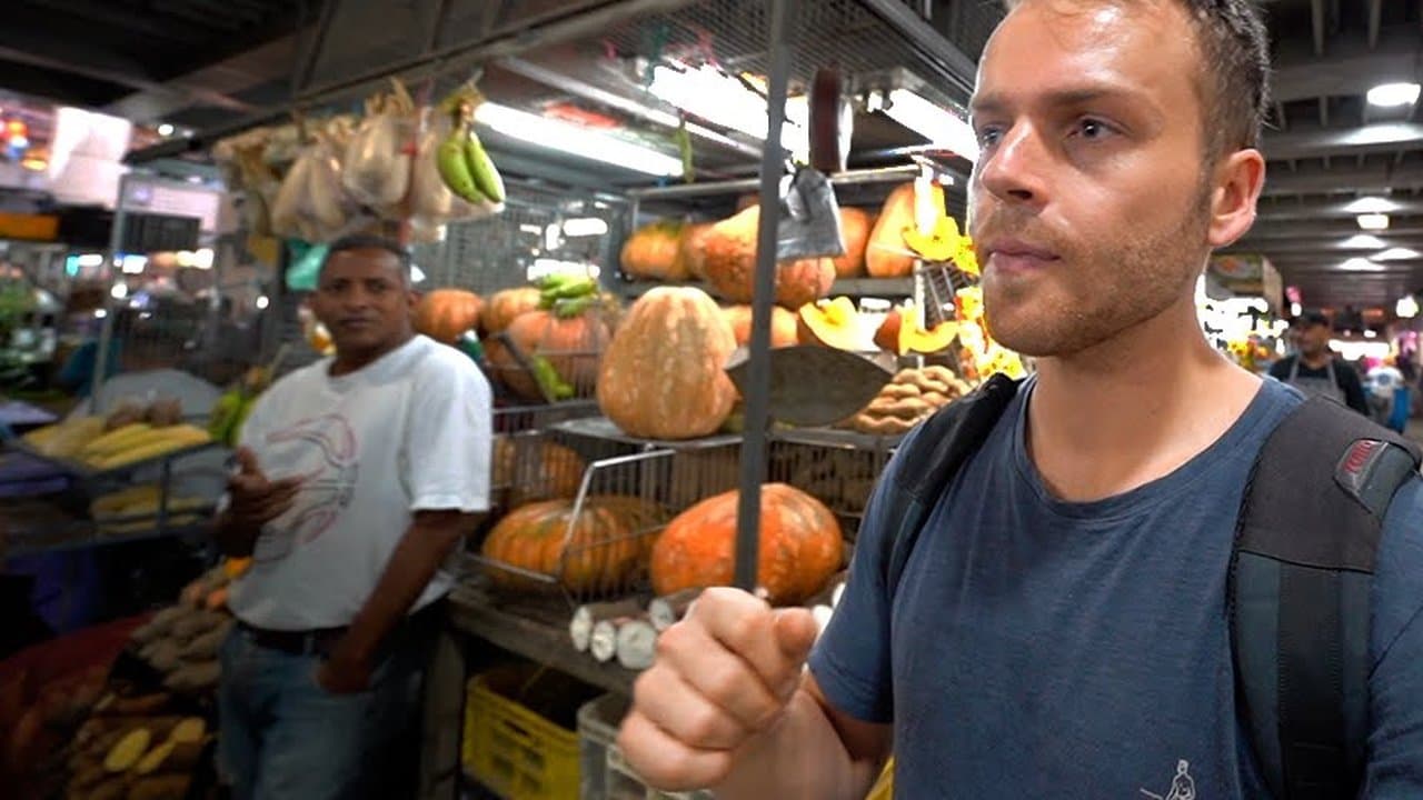 A young Nick Fisher with a backpack in a Caracas market hall, talking to the camera beside a stall piled with pumpkins, papayas and bananas while the stallholder looks on
