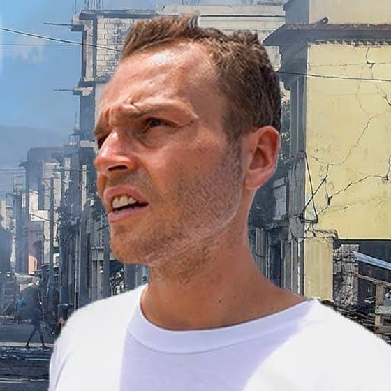 Nick Fisher in a white T-shirt, frowning and squinting into the sun on a smoky street in Port-au-Prince, Haiti, with cracked yellow concrete buildings behind him