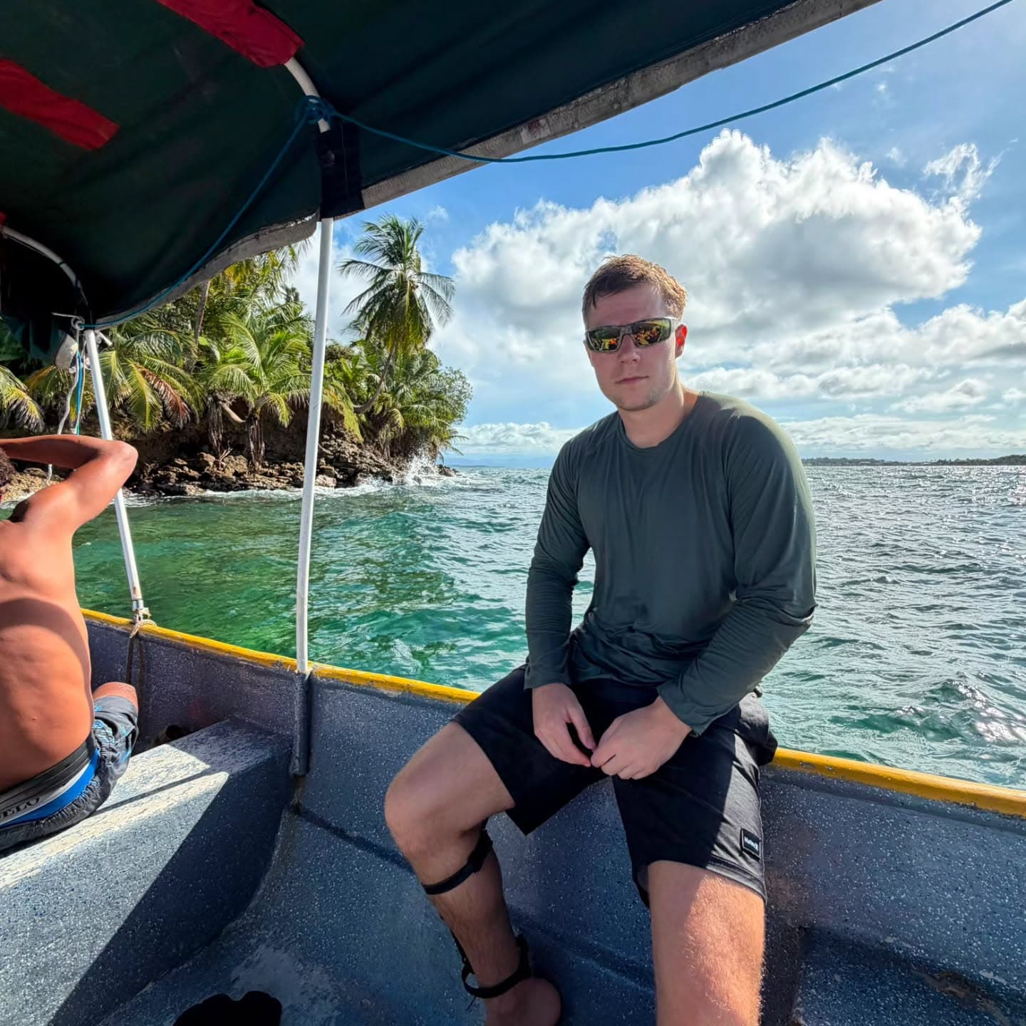 Kurt Caz sitting on the edge of a small boat off a palm-lined coastline, photo from his Instagram