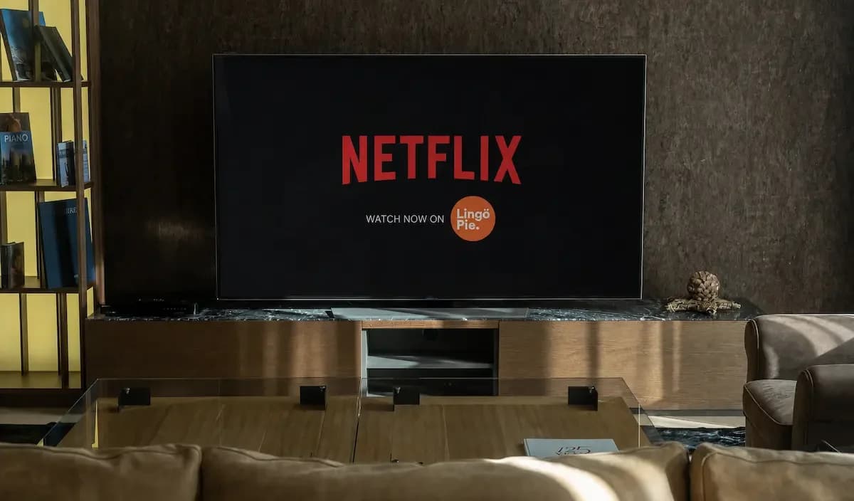 A big TV in a dim living room showing the red Netflix logo with an orange Lingopie badge and the words Watch now on, seen from behind a beige sofa