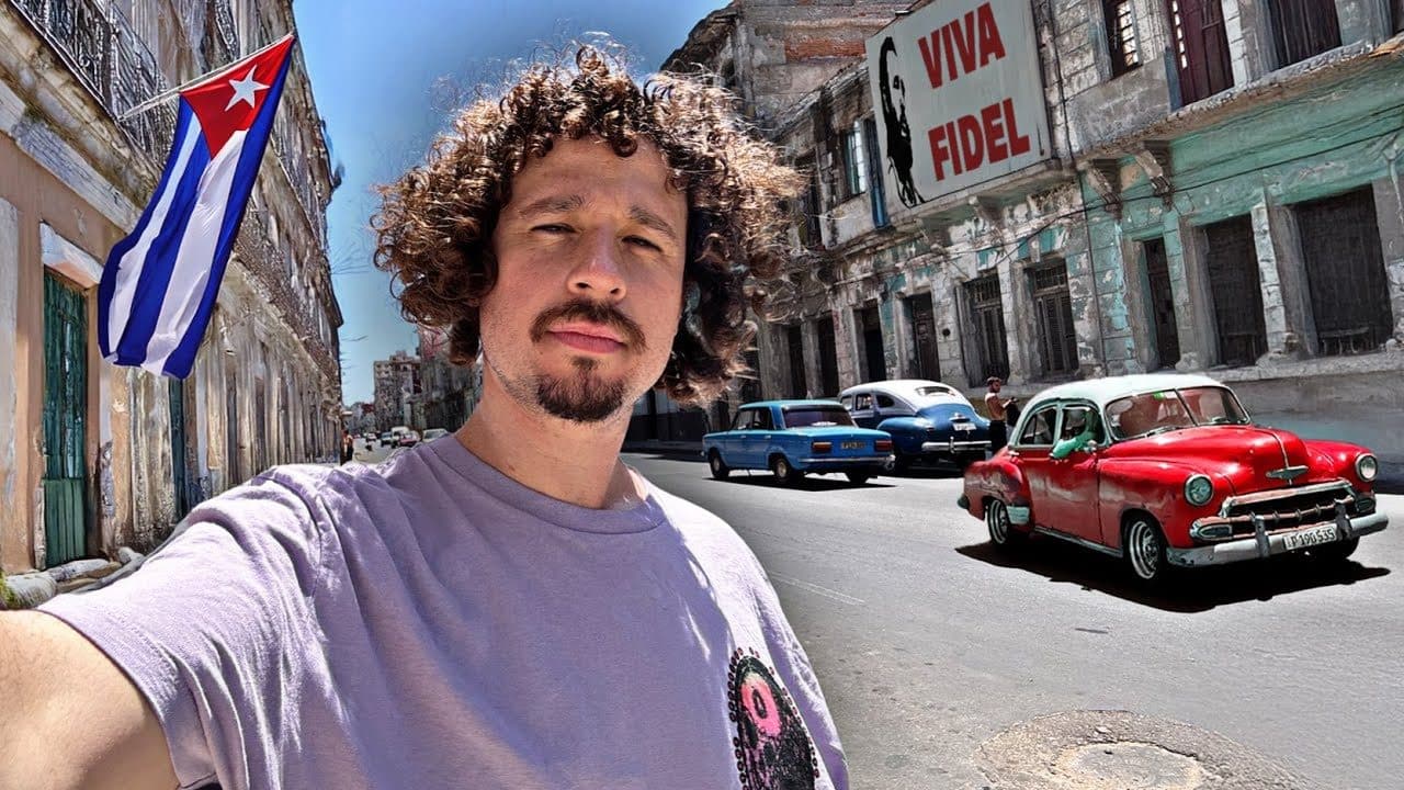 Luisito Comunica in a lilac T-shirt taking a selfie on a crumbling street in Cuba, with a Cuban flag on a balcony, a Viva Fidel sign on the wall and a red 1950s car driving past