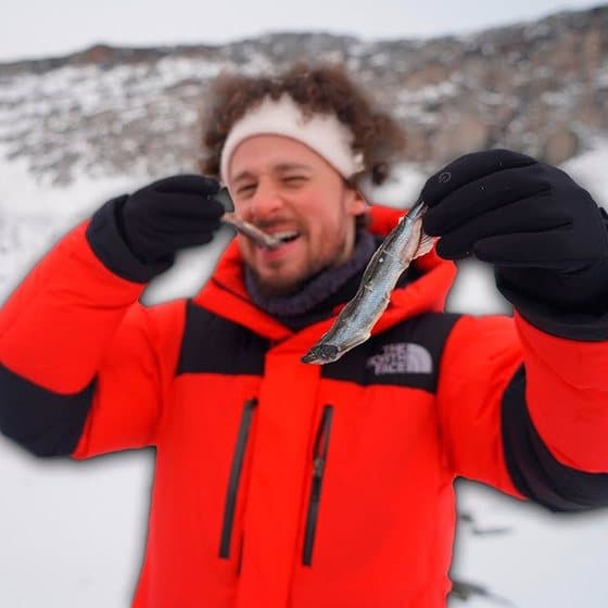Luisito Comunica on a snowy hillside in Greenland in a red down jacket, white headband and black gloves, biting one small dried fish and holding a second one out to the camera