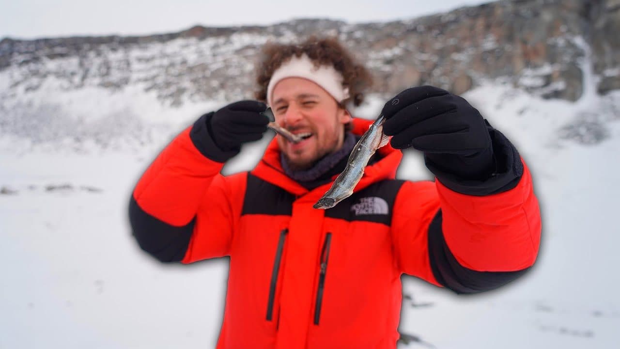 Luisito Comunica squinting with a small dried fish between his teeth and another held up in a gloved hand, in a red North Face jacket against snow and grey rock in Greenland
