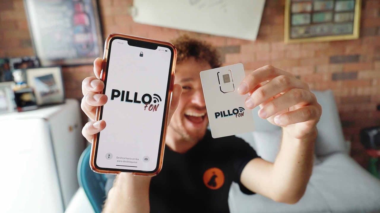 Luisito Comunica laughing as he holds up a phone showing the PilloFon logo, with PILLO as the network name in the corner of the screen, and a PilloFon SIM card in his other hand