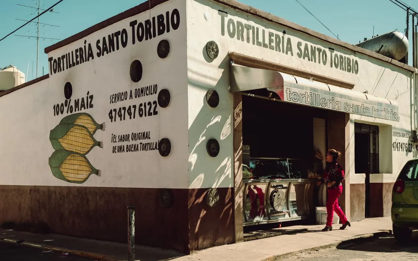 Tortillería Santo Toribio on a sunny corner: a white wall painted with ears of corn and the words 100% maíz, as a woman in red trousers walks into the shop