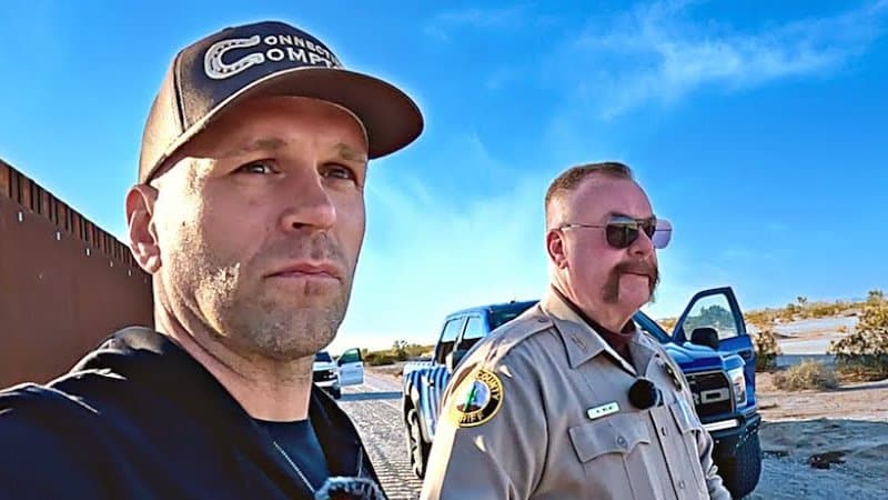 Peter Santenello in a cap and dark hoodie next to the rust-brown steel border wall in the Arizona desert, with a mustached sheriff in a tan uniform and sunglasses standing behind him by a pickup truck