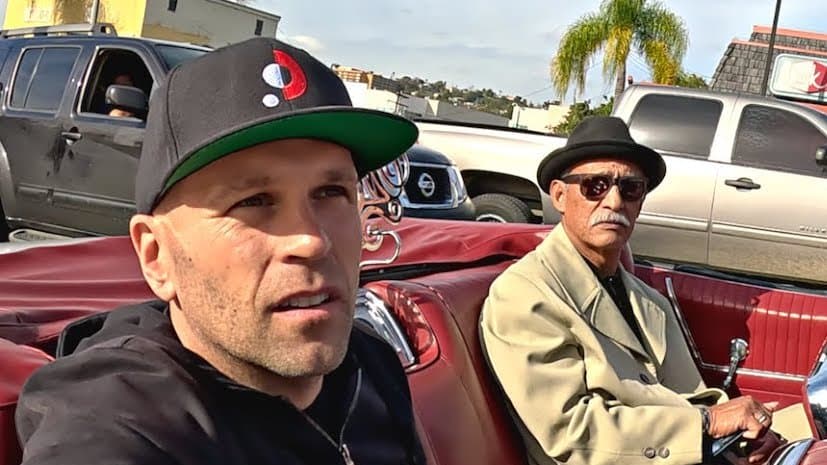 Peter Santenello in a black cap riding in the back of a red convertible lowrider in East LA, beside an older Chicano man in a black hat, tinted glasses and a cream overcoat