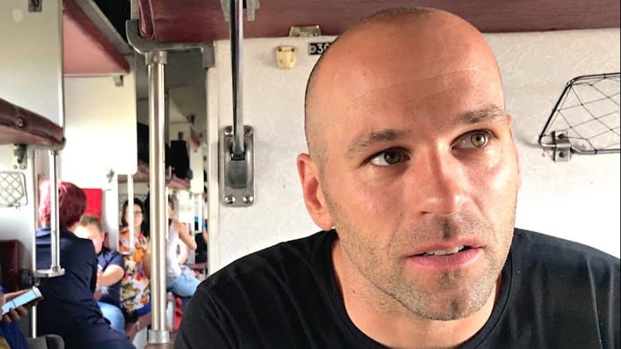Peter Santenello in a black T-shirt at the window of an open Ukrainian sleeper carriage, with passengers on the lower bunks behind him, on the overnight train to the village