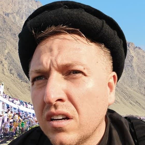 Simon Wilson in a black Chitrali-style wool cap, squinting at the camera with a doubtful look under a bright sky in Pakistan, dry brown mountains and a crowded train behind him