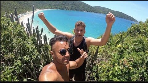 Timmy Karter cheering with both arms raised on a cactus-covered hill above the turquoise bay of Arraial do Cabo, Brazil, with a friend in sunglasses in front of him