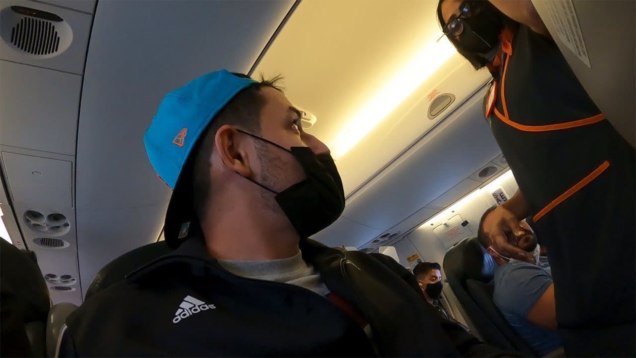 Volpe in a turquoise baseball cap worn backwards and a black face mask, looking up at a flight attendant in a dark uniform with orange trim in the aisle of a plane, other masked passengers behind him