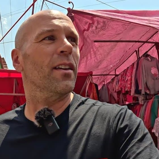 Peter Santenello, bald and stubbled, in a black T-shirt with a clip-on microphone, looking up at a Mexico City street market where children's clothes hang under pink and red awnings