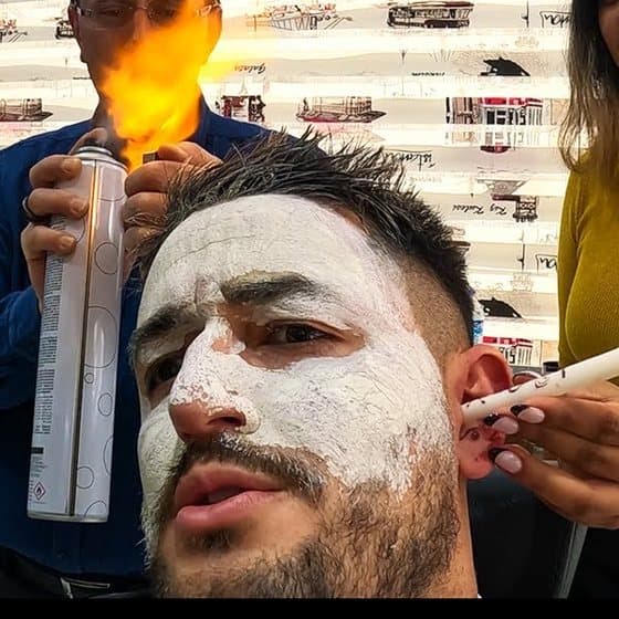 Volpe with a white clay mask on his face in an Istanbul barbershop, wincing while the barber sends a flame over his hair from a spray can and an assistant in a yellow jumper holds a lit ear candle to his ear